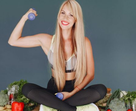 Woman lifting a small dumbbell while surrounded by fresh vegetables, embodying health and fitness.