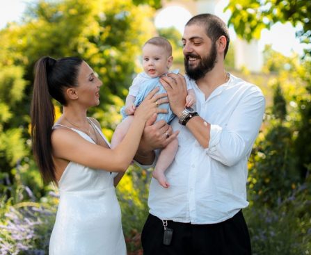 Mother and father joyfully hold their baby in a vibrant outdoor garden setting.